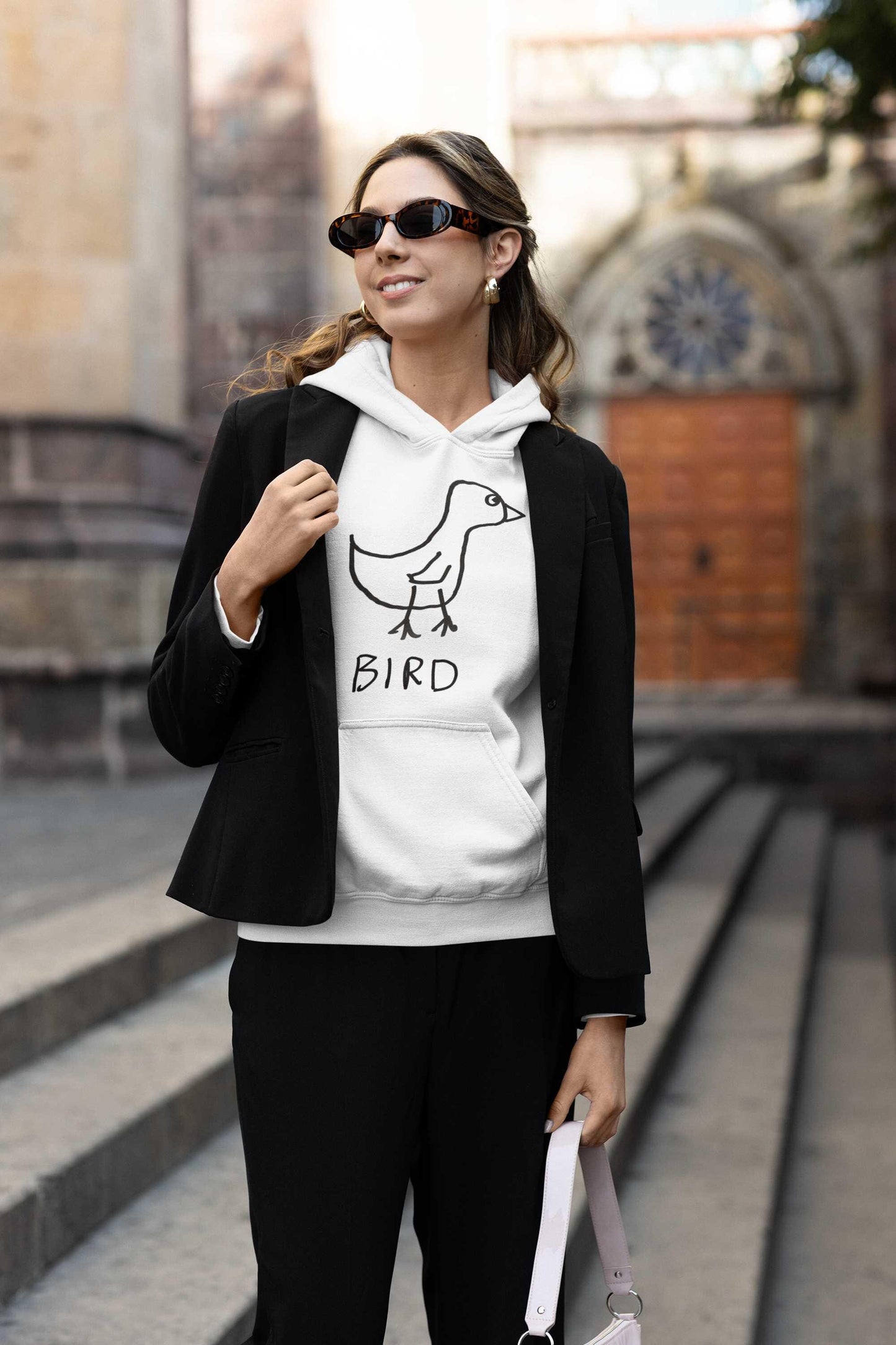 fashionable young woman wearing a jacket over a white hooded pullover sweatshirt featuring a crude marker drawing of a bird with the word "Bird" underneath