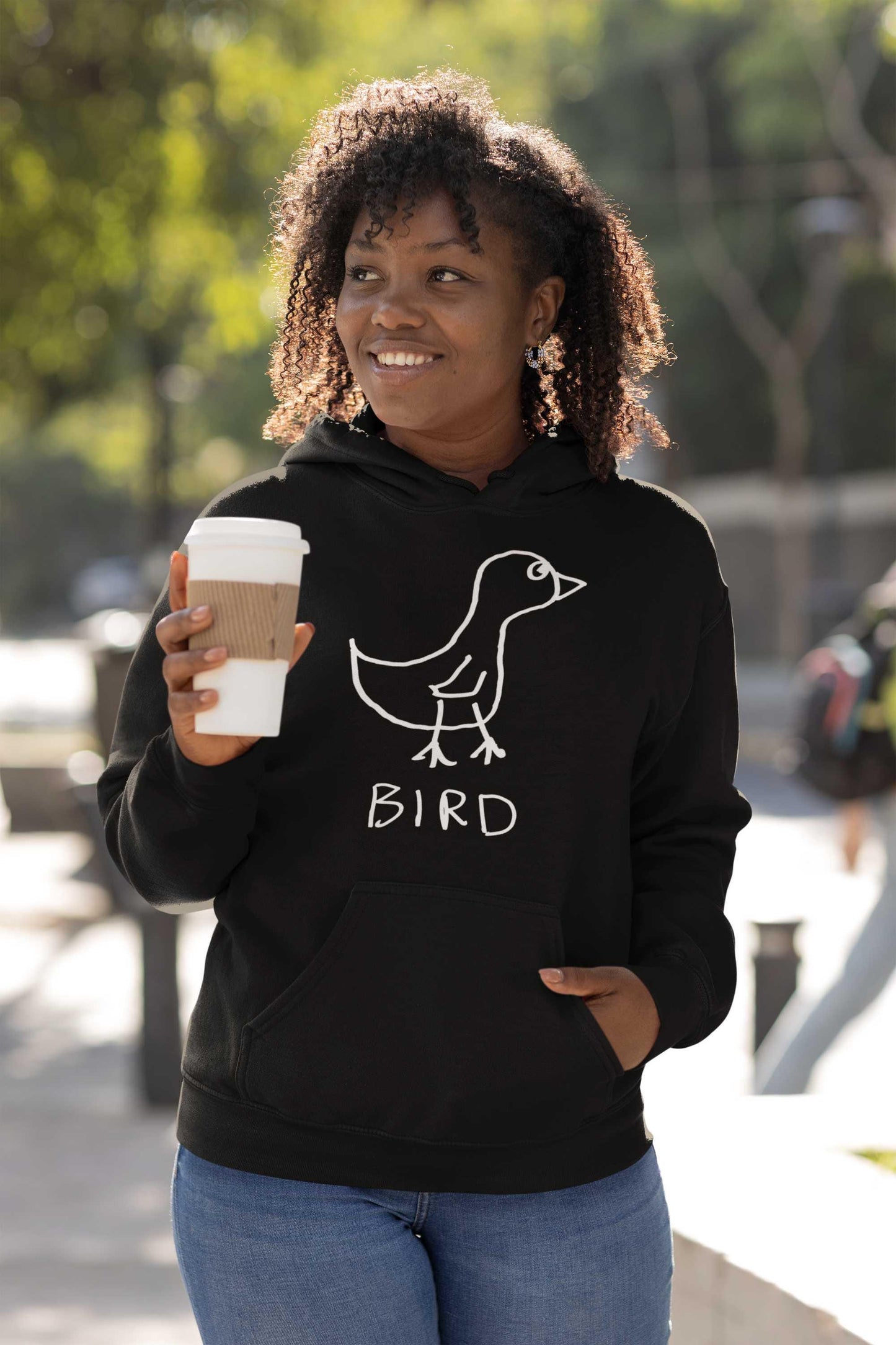 woman outdoors holding a coffee wearing a black hooded pullover sweatshirt featuring a crude marker drawing of a bird with the word "Bird" underneath