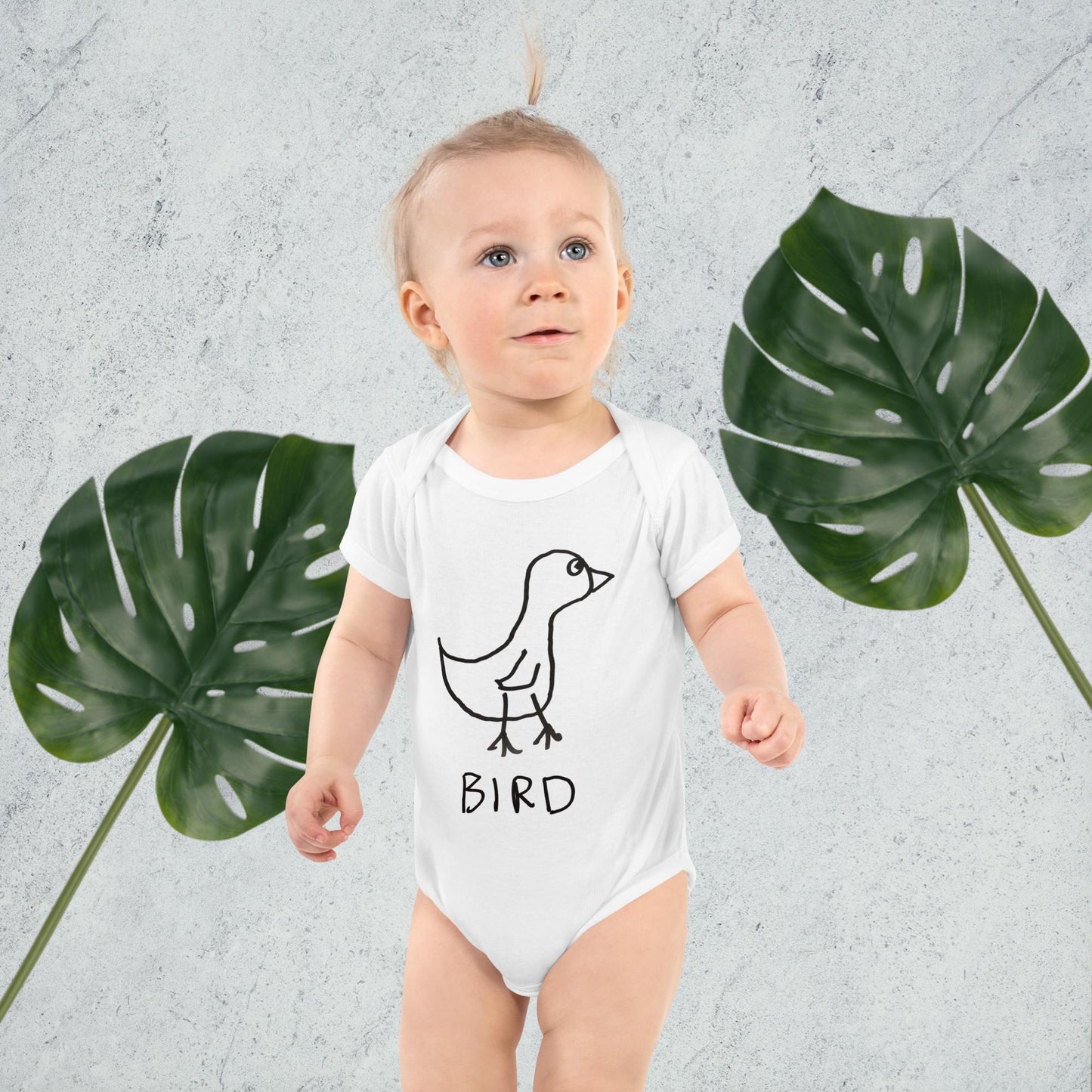 a cute blonde baby wearing a white baby onesie featuring a crude marker drawing of a bird with the word "Bird" underneath standing against a gray background with green leaves.