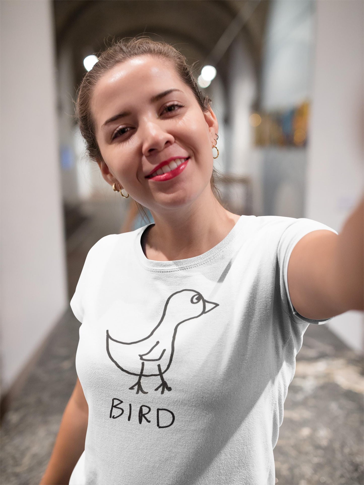A smiling young woman wearing a fitted t-shirt featuring a crudely drawn black marker drawing of a bird with the word "Bird" underneath