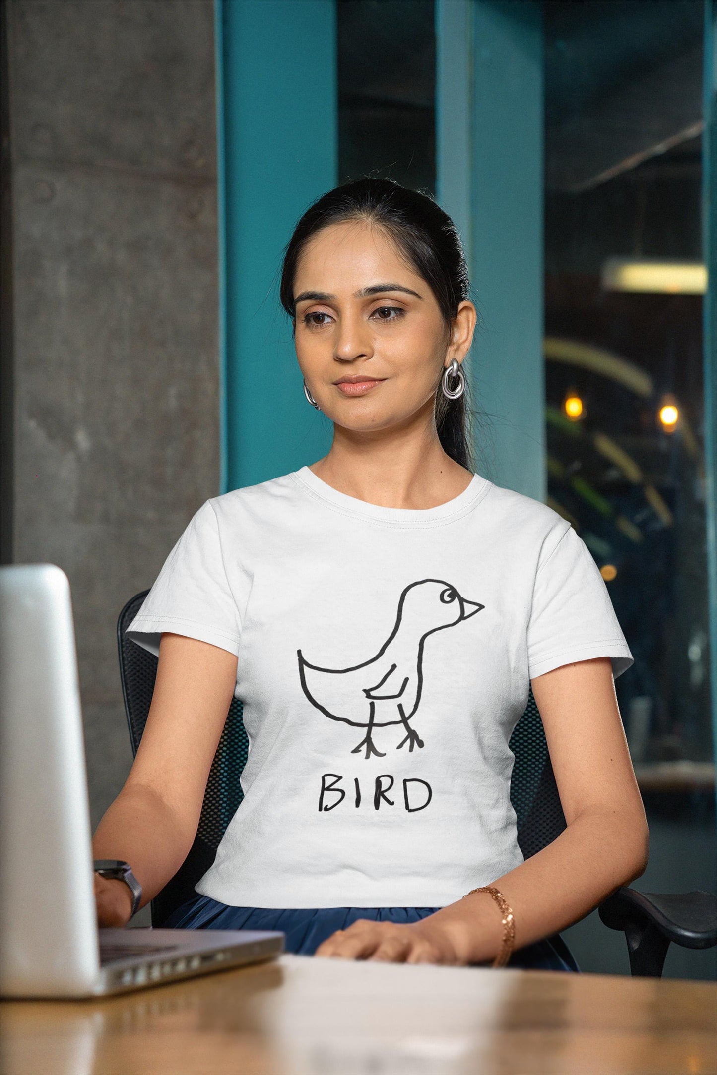 A young woman sitting at her laptop computer, wearing a white fitted t-shirt featuring a crudely drawn black marker drawing of a bird with the word "Bird" underneath