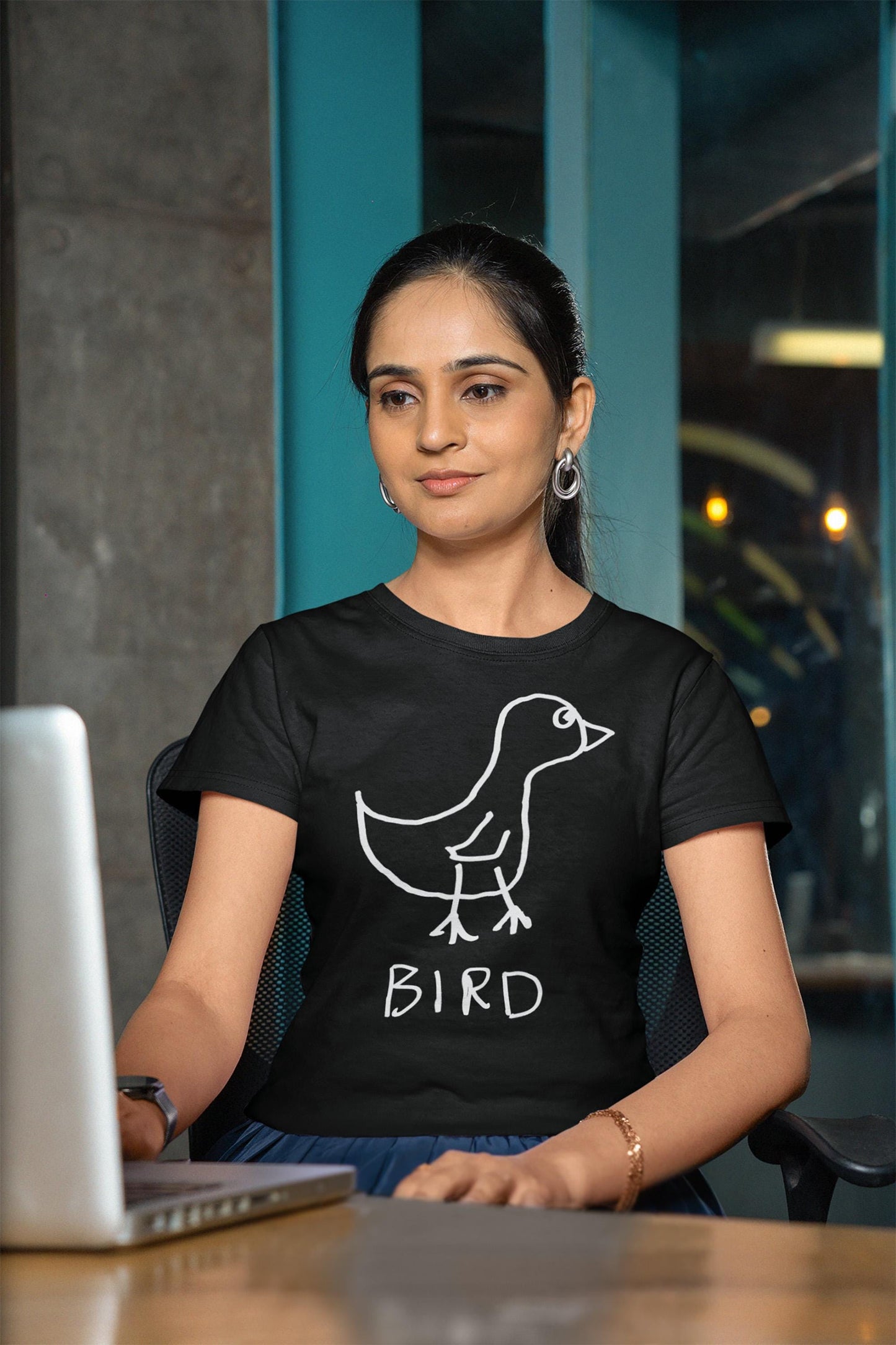 A young woman sitting at her laptop computer, wearing a black fitted t-shirt featuring a crude marker drawing of a bird with the word "Bird" underneath