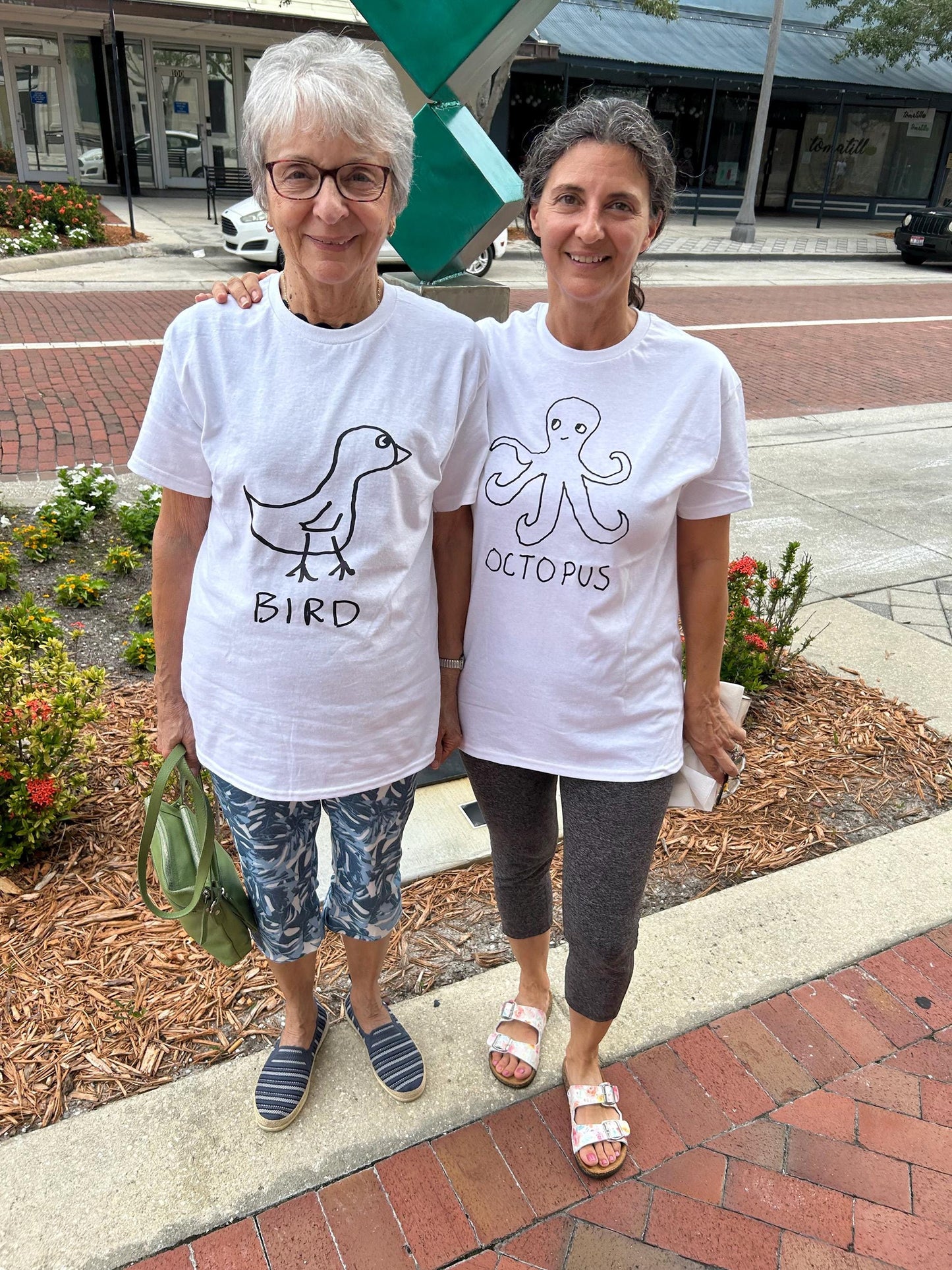 Two women wearing white t-shirts featuring a crude marker drawing of a bird with the word "Bird" underneath, standing on a sidewalk.