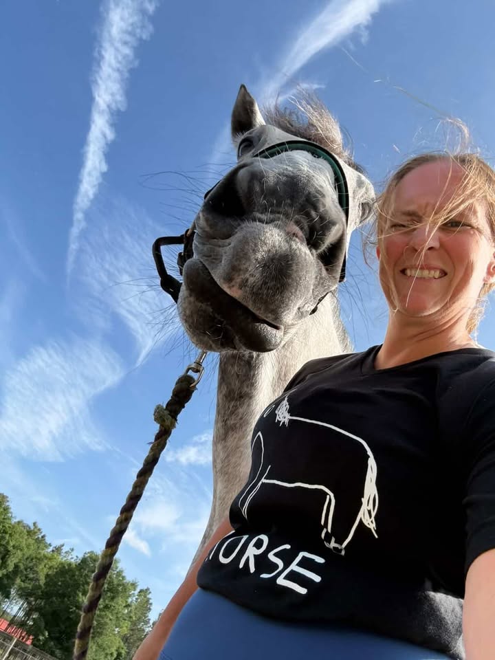 Woman wearing a black t-shirt with a lo-fi hand-drawn horse design, standing next to a horse against a blue sky.