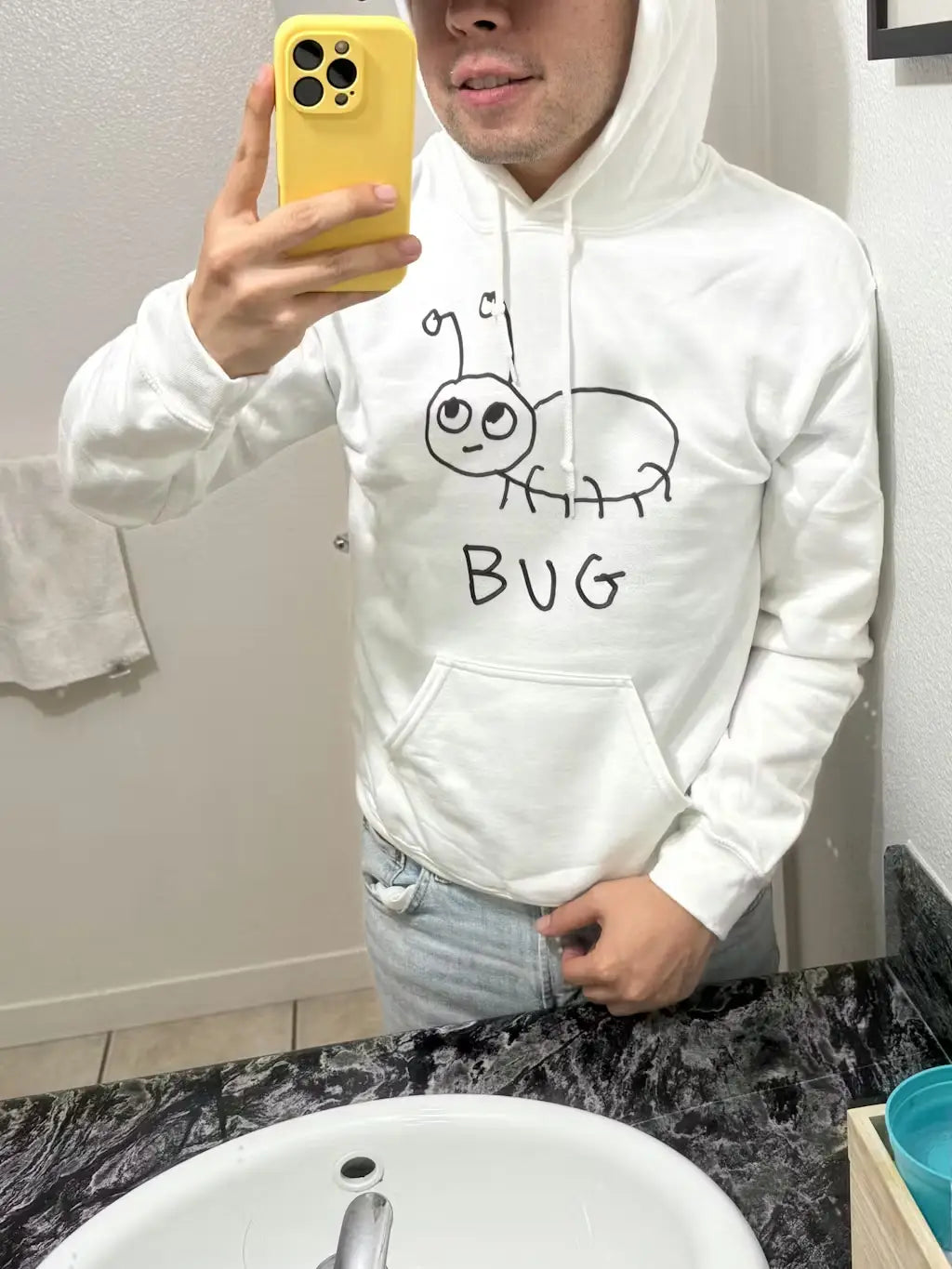 A young man taking a selfie, posing in the mirror, wearing a white hoodie sweatshirt with a lo-fi hand-drawn bug graphic and text "BUG"