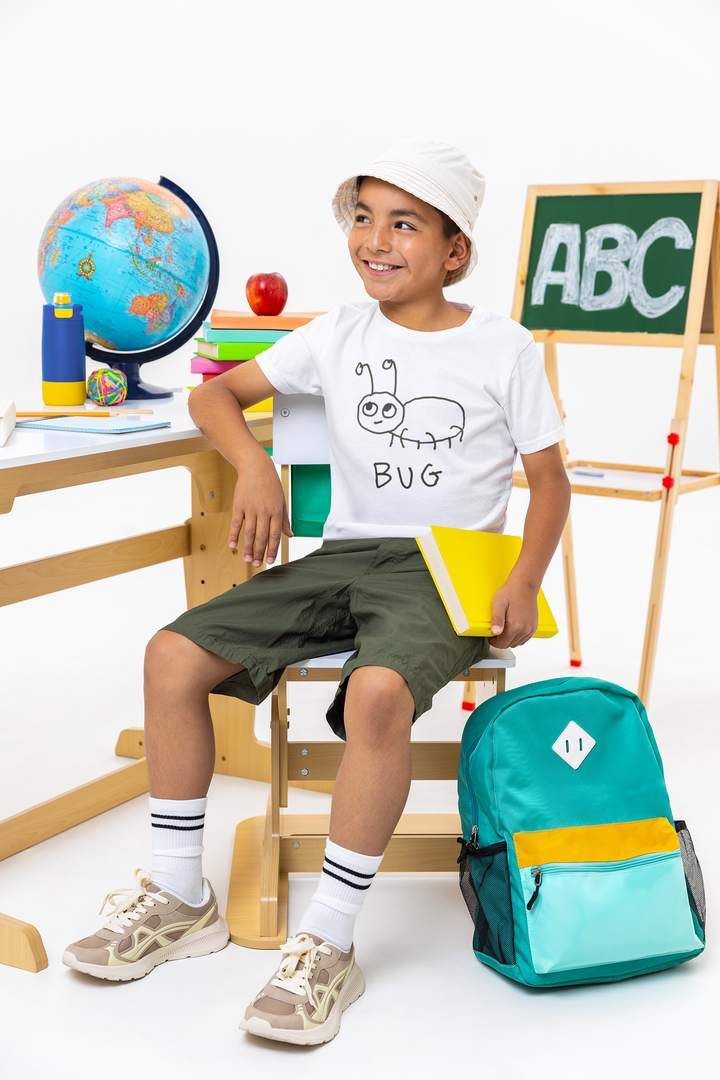 Child wearing a white t-shirt with simple hand drawn bug illustration and text, sitting on a chair with educational items and a backpack in the background