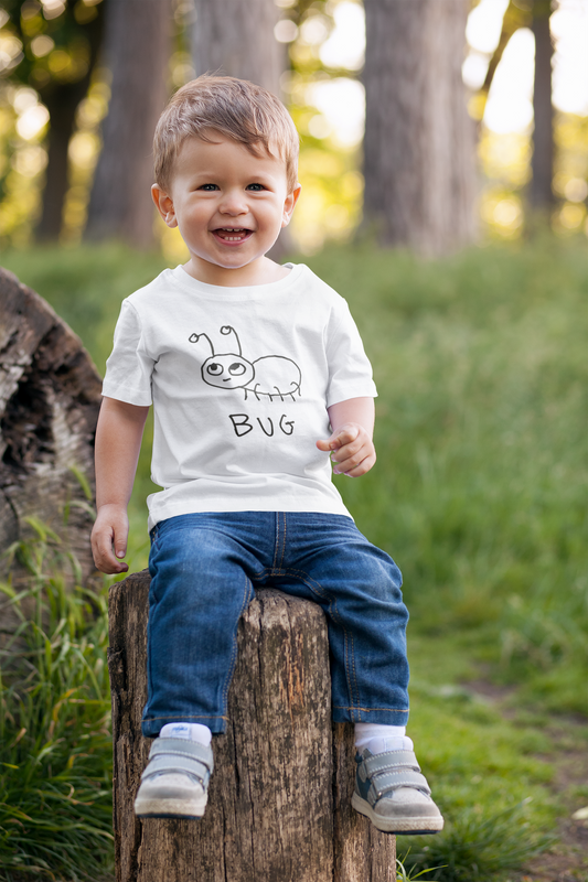 Toddler wearing a white t-shirt with a simple hand drawn 'bug' graphic sitting on a log in a forest.