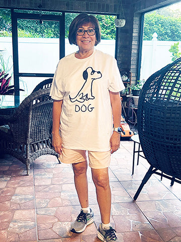 a lady standing on her back screened porch, wearing a white t-shirt featuring a lo-fi hand-drawn dog graphic and text. 