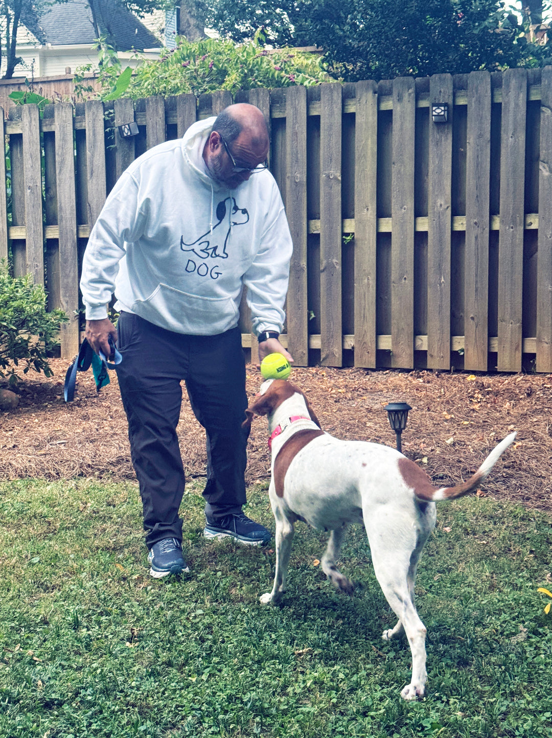 Man playing with a dog in a backyard, wearing a white hoodie with a quirky minimalist lofi DOG drawing,