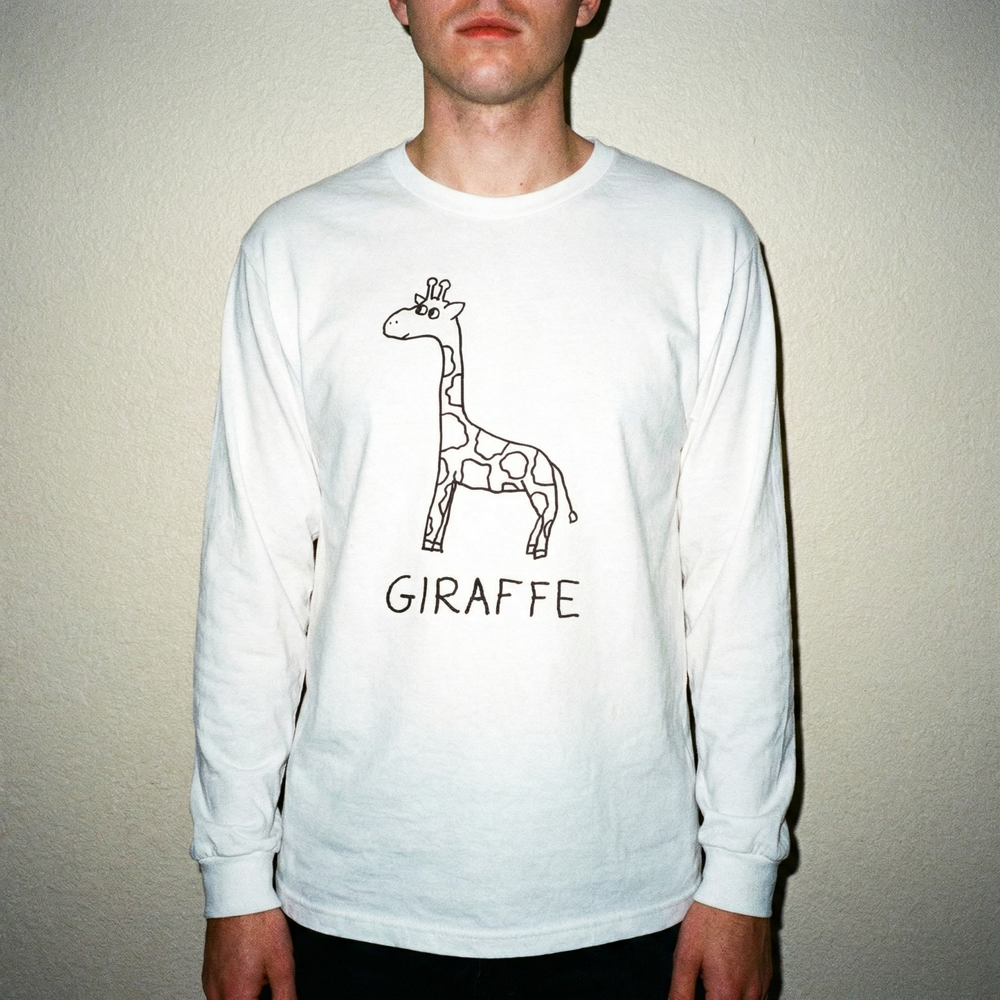 Model wearing a white long-sleeve tee shirt with a hand drawn giraffe graphic and text, beige background, flash 