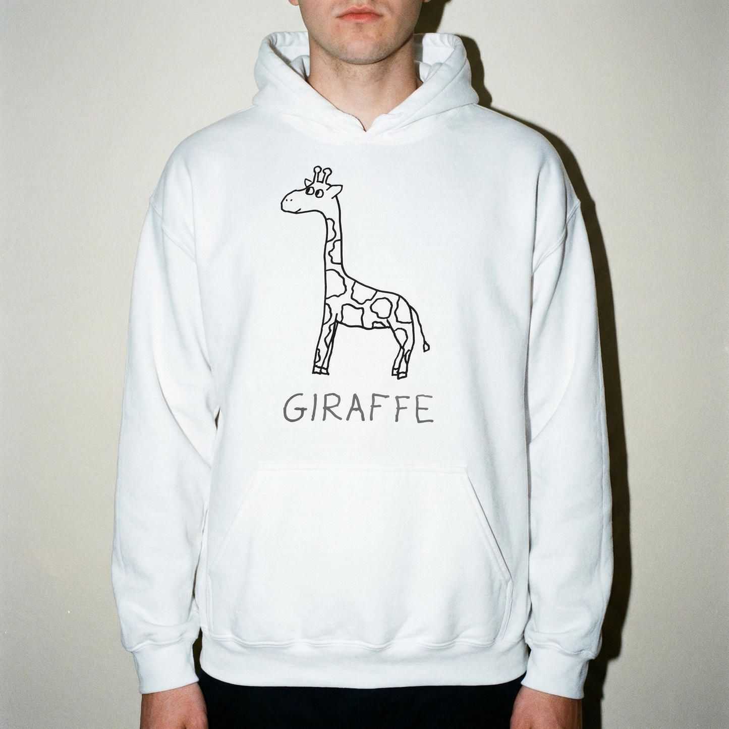Young man in studio setting wearing a white hoodie with a quirky minimalist lofi giraffe drawing. 