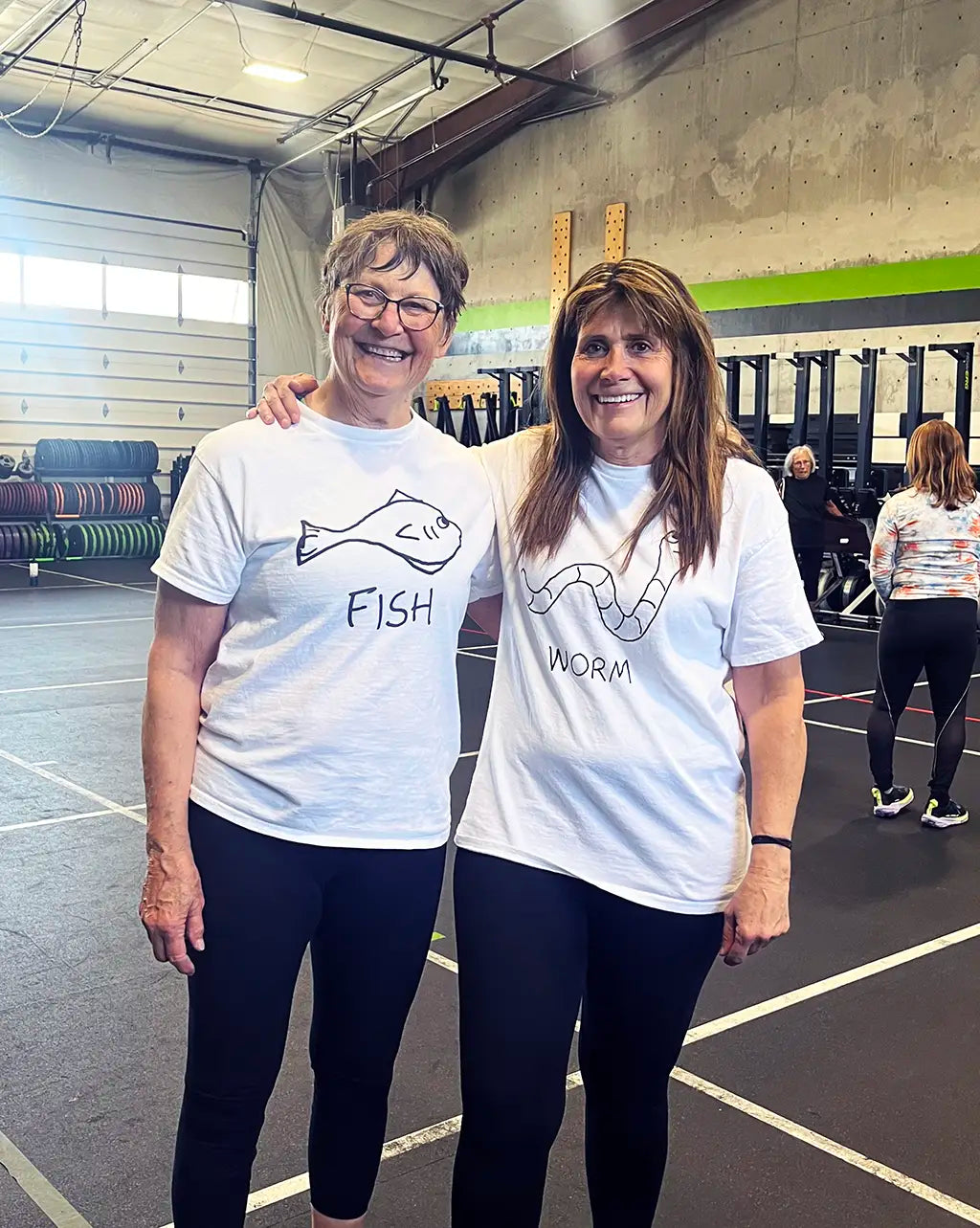 Two women at a gym, standing arm in arm, both wearing white t-shirts with lo-fi hand-drawn animal graphics and text . One fish, one worm.