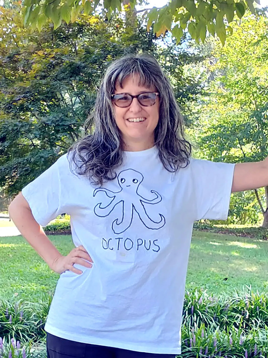 A woman in a park wearing sunglasses and a white t-shirt with a lo-fi hand-drawn octopus graphic and text. 