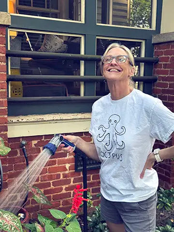 Woman wearing a white t-shirt with a lo-fi hand-drawn octopus graphic and text,  holding a garden hose in front of a brick building with windows.
