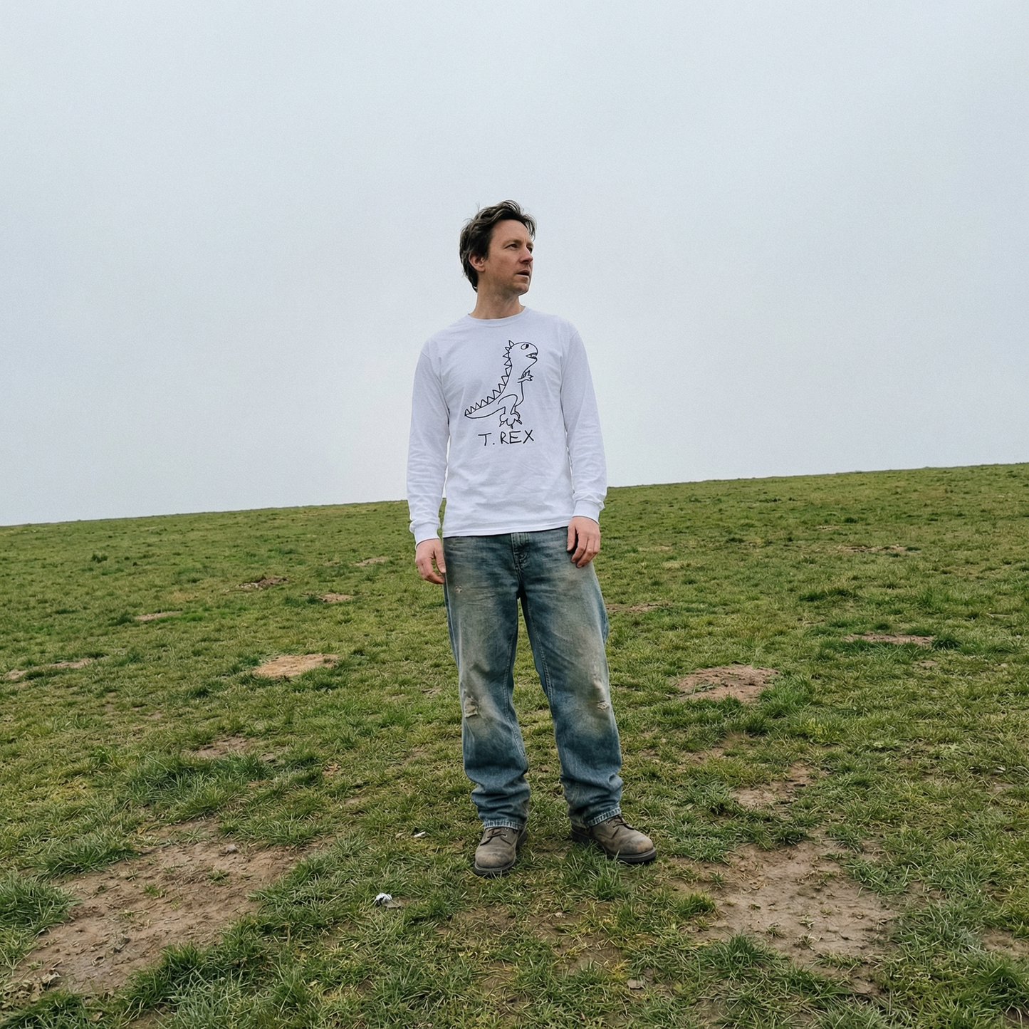 Model in a T-Rex long sleeve tee alone in a flat grassy field under a grey sky