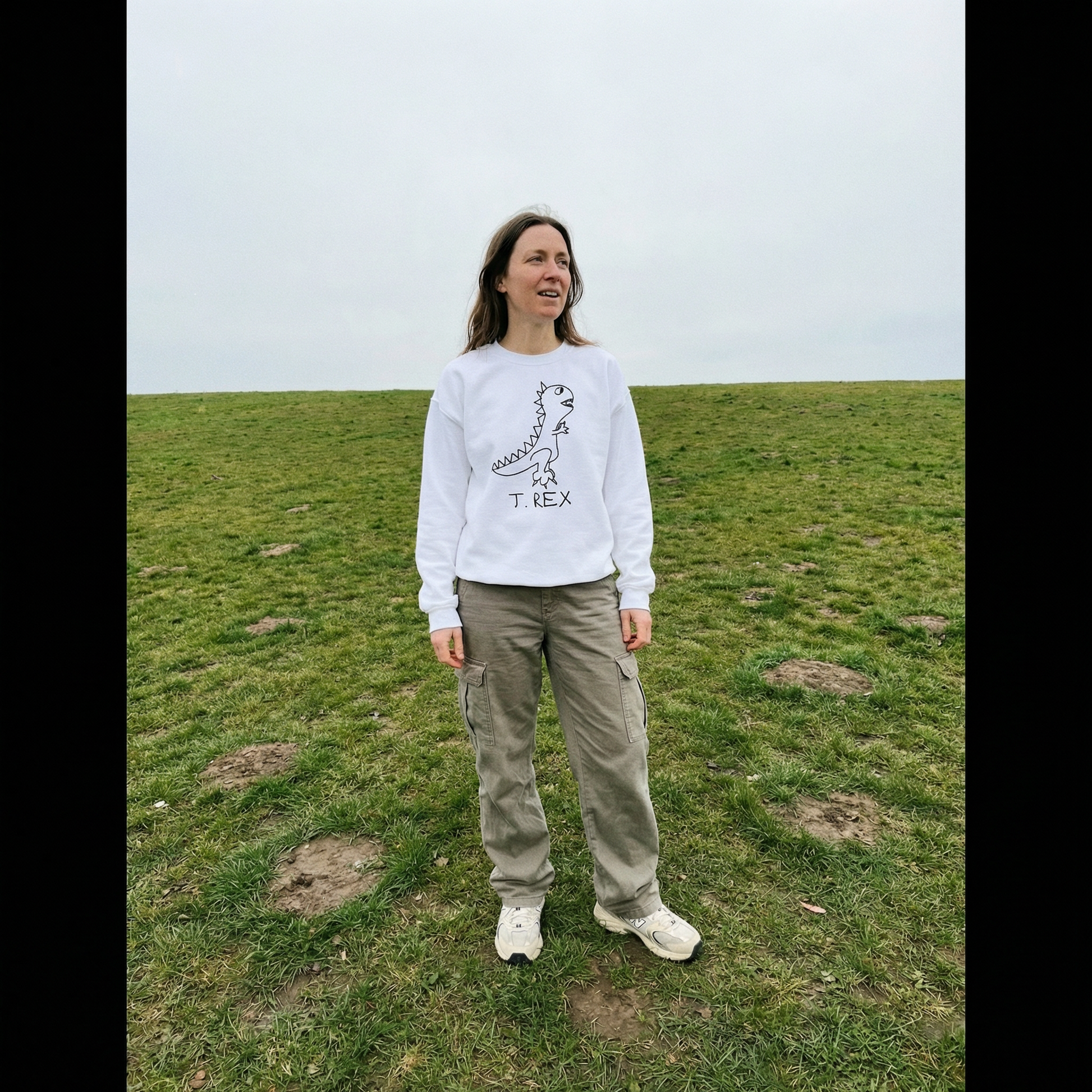 Model in a T-Rex sweatshirt standing wide-legged in a grassy field, overcast and dramatic