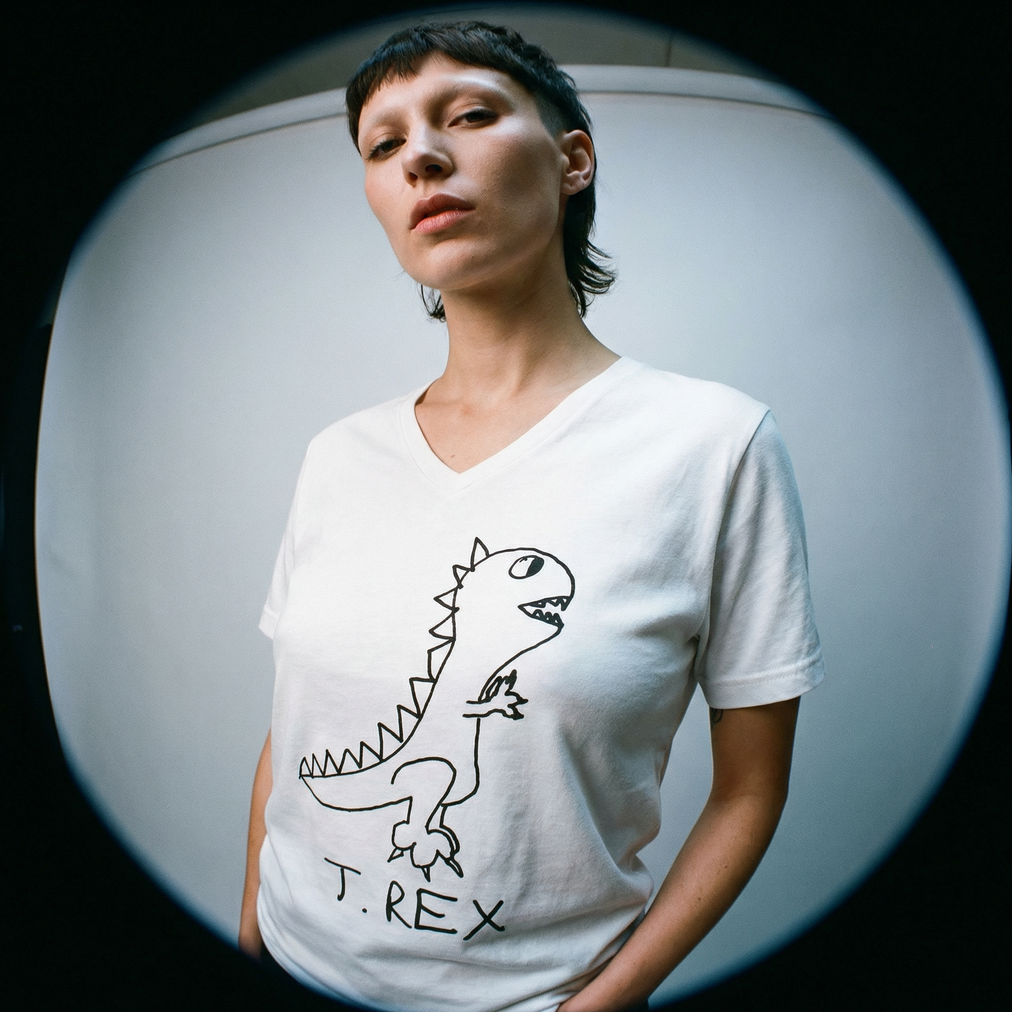 Model in T-Rex graphic v-neck tee in fisheye lens against a gray wall.