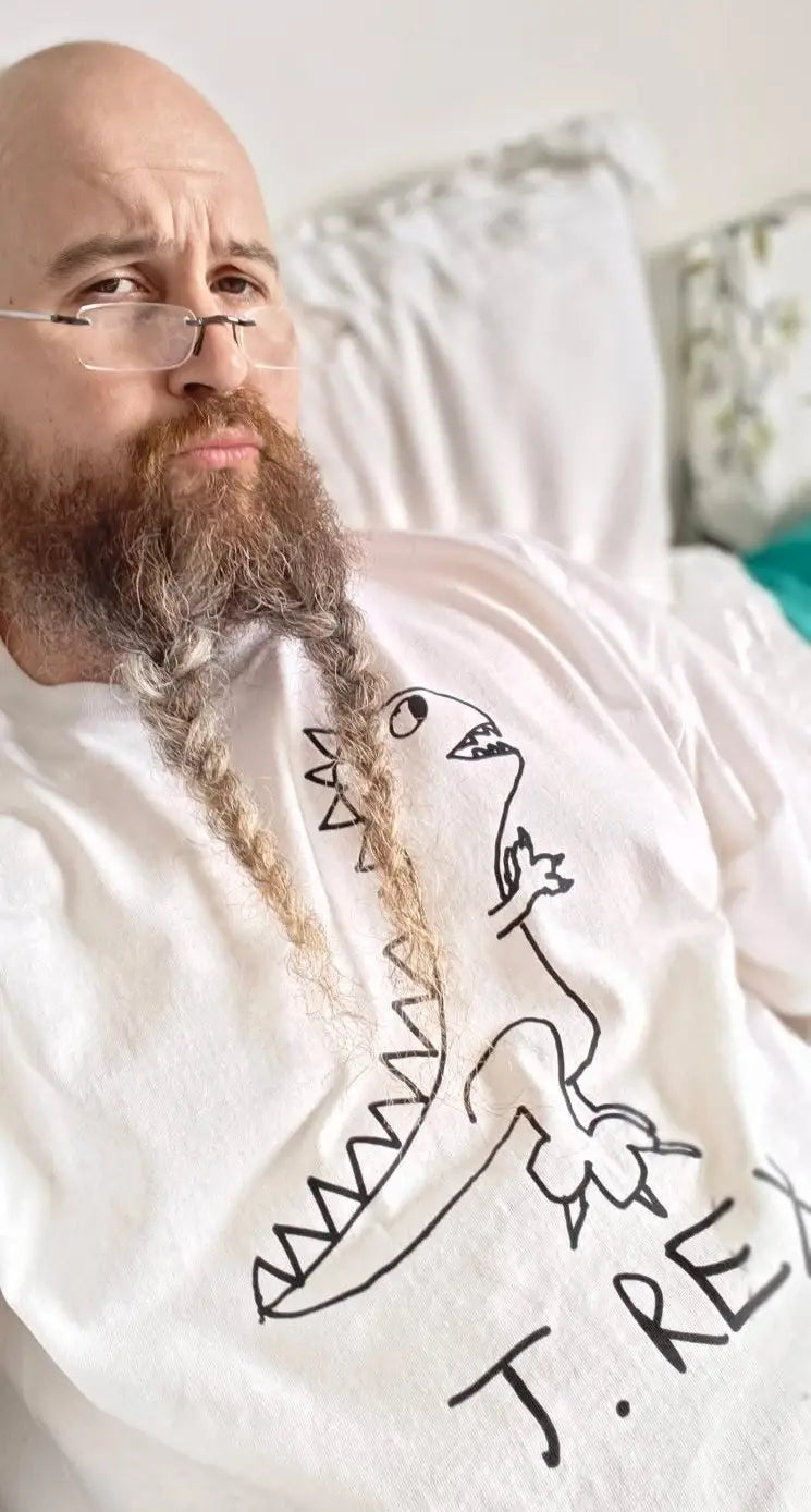 Man with braided beard wearing a white t-shirt with a lo-fi hand drawn graphic of a T. Rex dinosaur, sitting on a couch. Tyrannosaurus Rex.