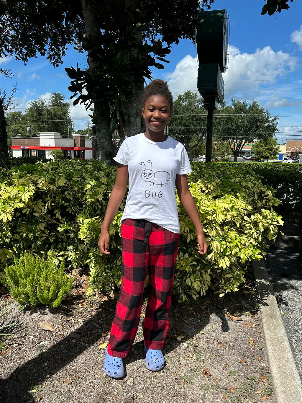 Young black woman wearing a white t-shirt featuring a lo-fi hand-drawn illustration of a bug, and red plaid pants standing outdoors with trees and sky in the background.