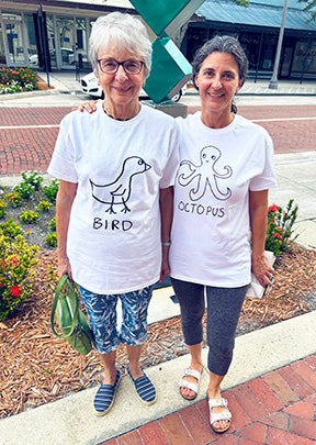 Two ladies standing on the sidewalk wearing white t-shirts with lo-fi hand-drawn animal graphics and text. One bird, one octopus. 