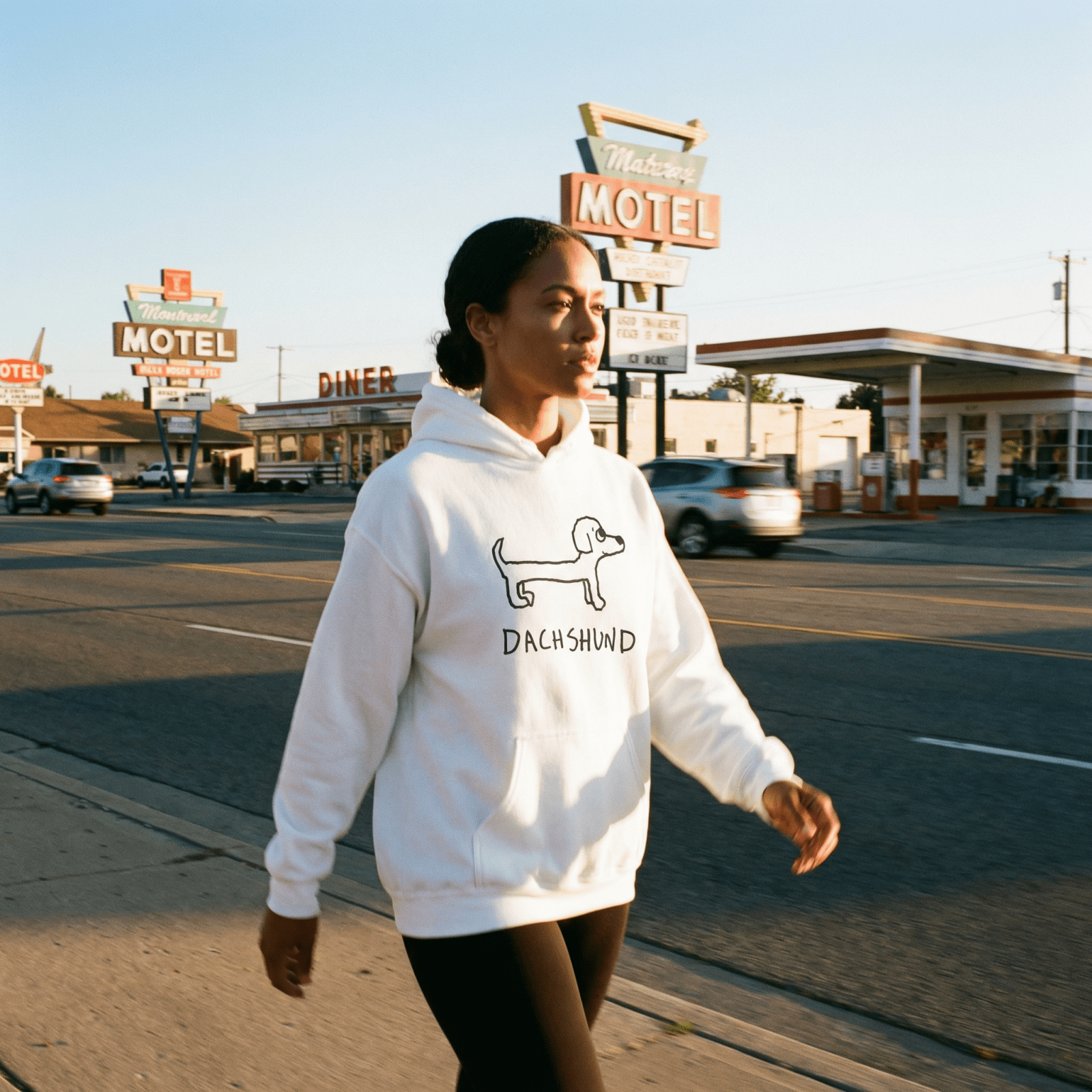 Woman walking past a diner and motel signs at golden hour wearing a lo-fi Dachshund Hoodie