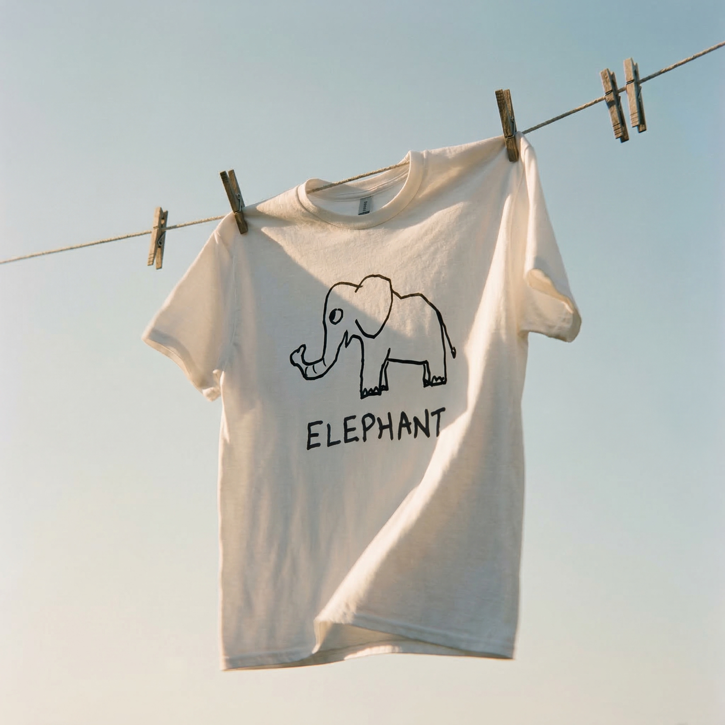 White t-shirt with an elephant graphic and 'ELEPHANT' text hanging on a clothesline against a clear sky.