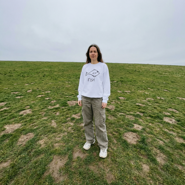 Person wearing a white crewneck sweatshirt with a  hand drawn fish graphic in a grassy field