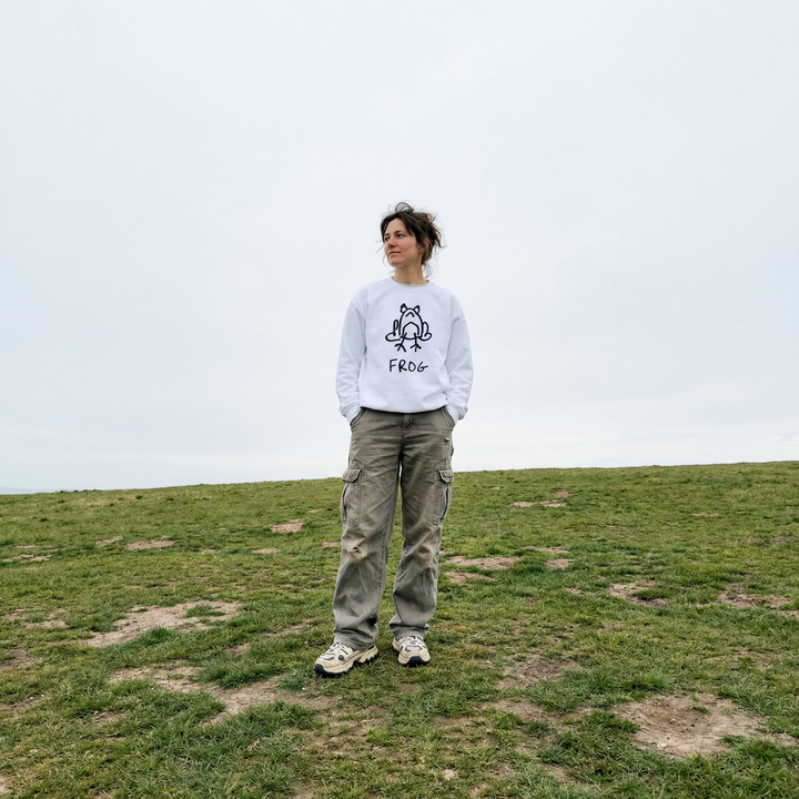Model in a frog sweatshirt standing in tall grass, overcast sky, easy slouch