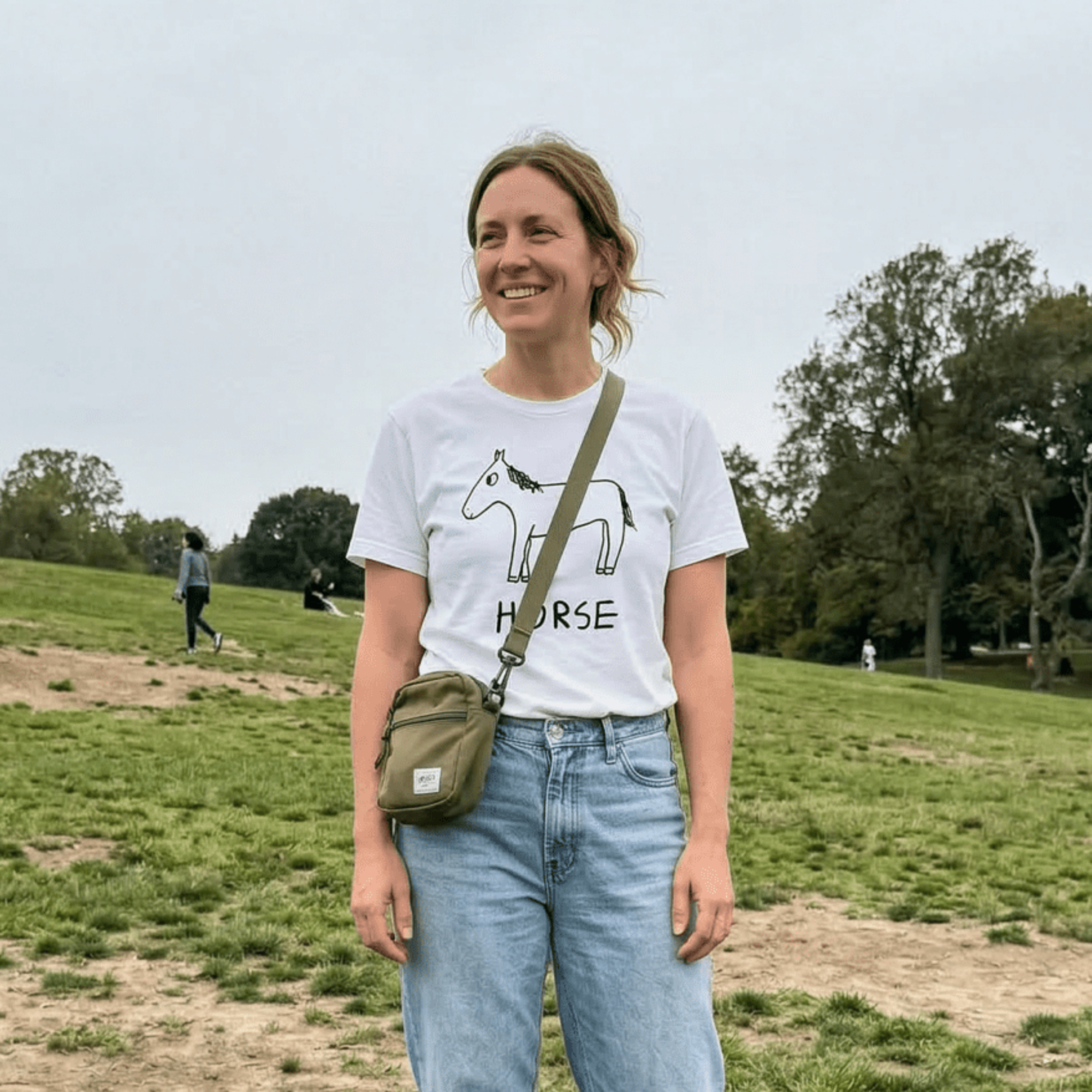 Woman with a crossbody bag standing in a park wearing a lo-fi Horse Classic Tee