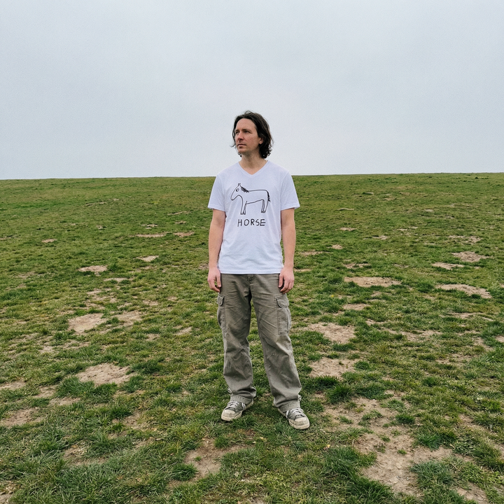 Model in horse v-neck tee in a wide rolling field, distant treeline behind them