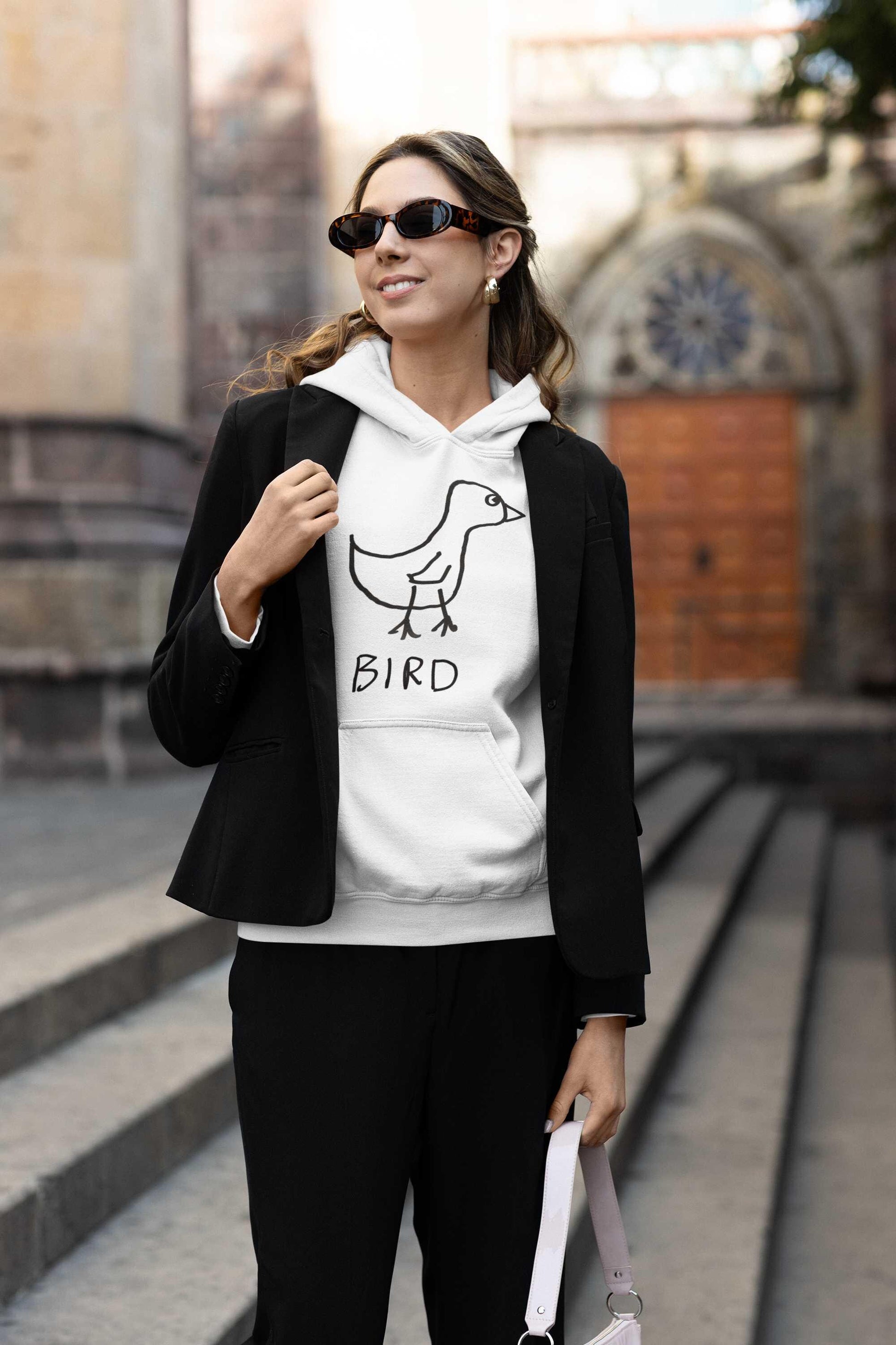 fashionable young woman wearing a jacket over a white hooded pullover sweatshirt featuring a crude marker drawing of a bird with the word "Bird" underneath