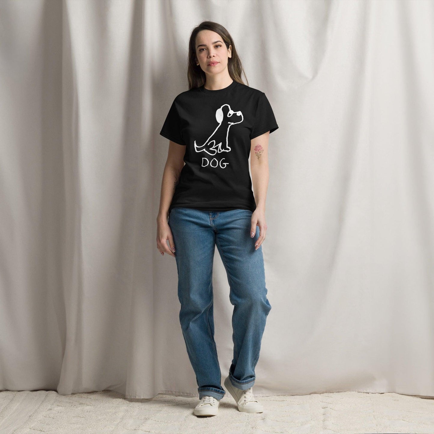 Pretty young woman standing against a white curtain background, wearing jeans and a black classic t-shirt  featuring a crude marker black and white marker drawing of a cute dog with the word "DOG" handwritten underneath