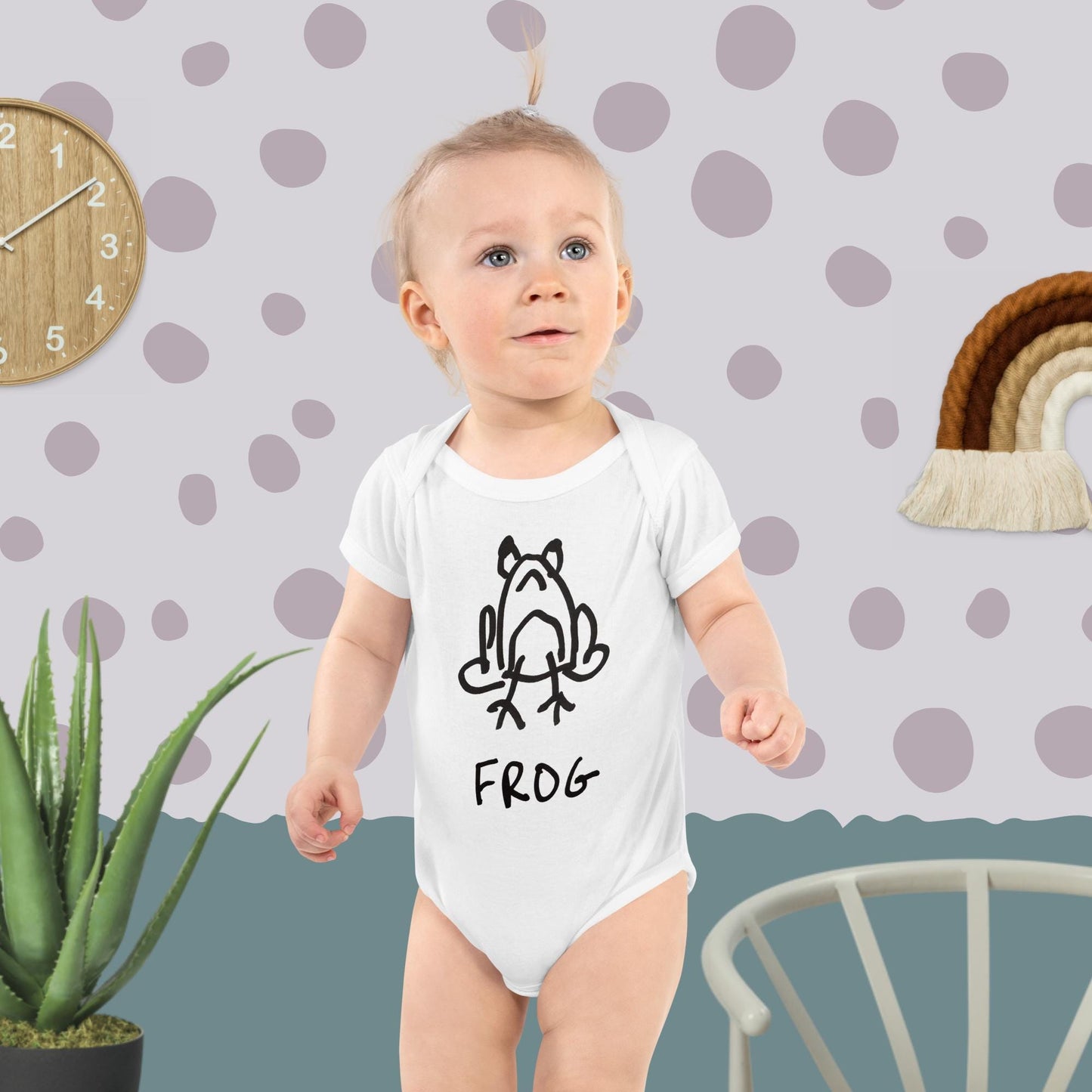 Baby wearing a white onesie featuring a crude but cute black and white marker marker drawing of a cute frog with the word “FROG” handwritten underneath standing against a polka dot wall.