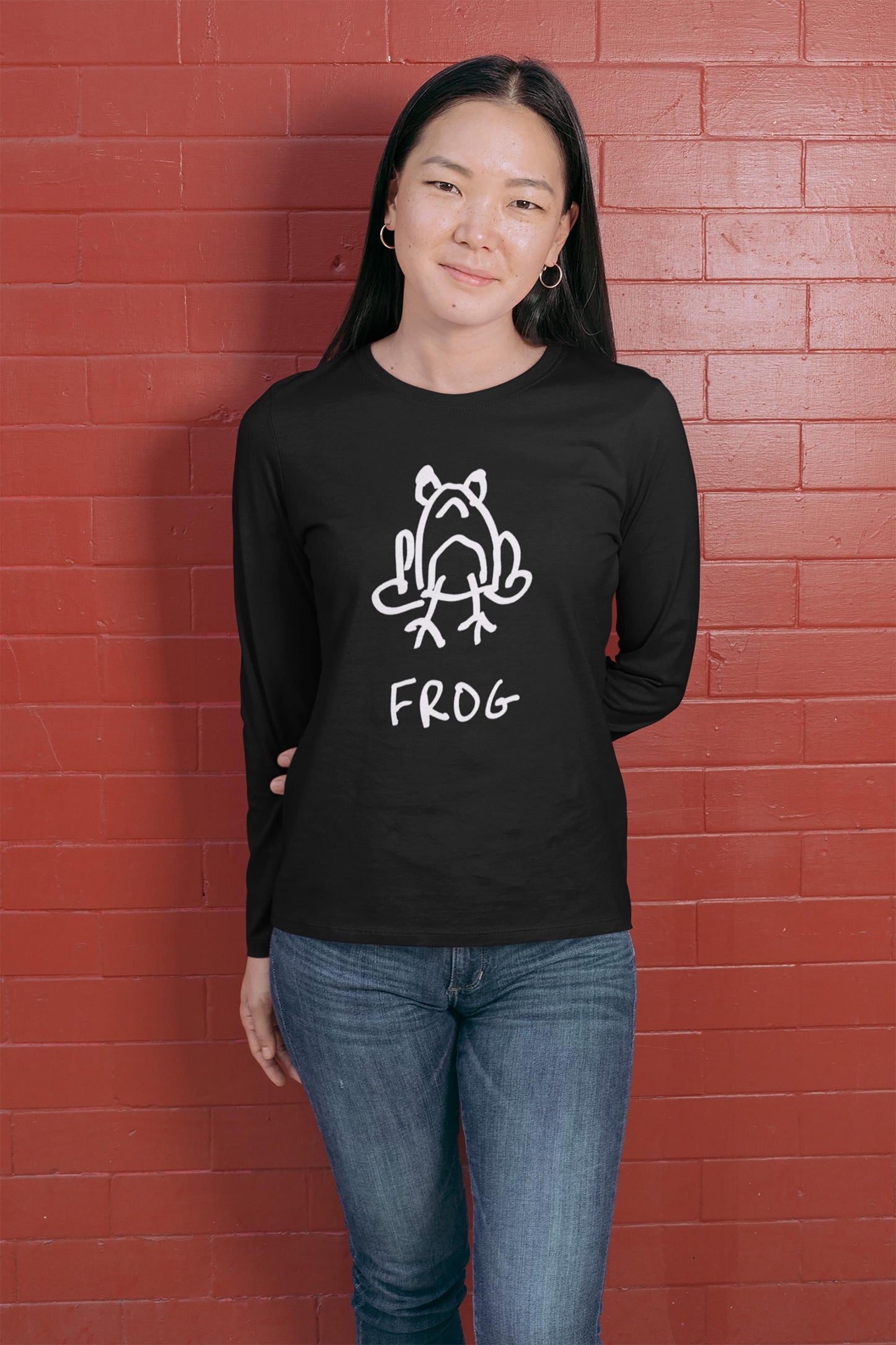 Smiling women wearing a black long-sleeve shirt Hip woman on a white background wearing a white long sleeved tee shirt featuring a crude but cute black and white marker marker drawing of a cute frog with the word “FROG” handwritten underneath, standing against a red brick wall.