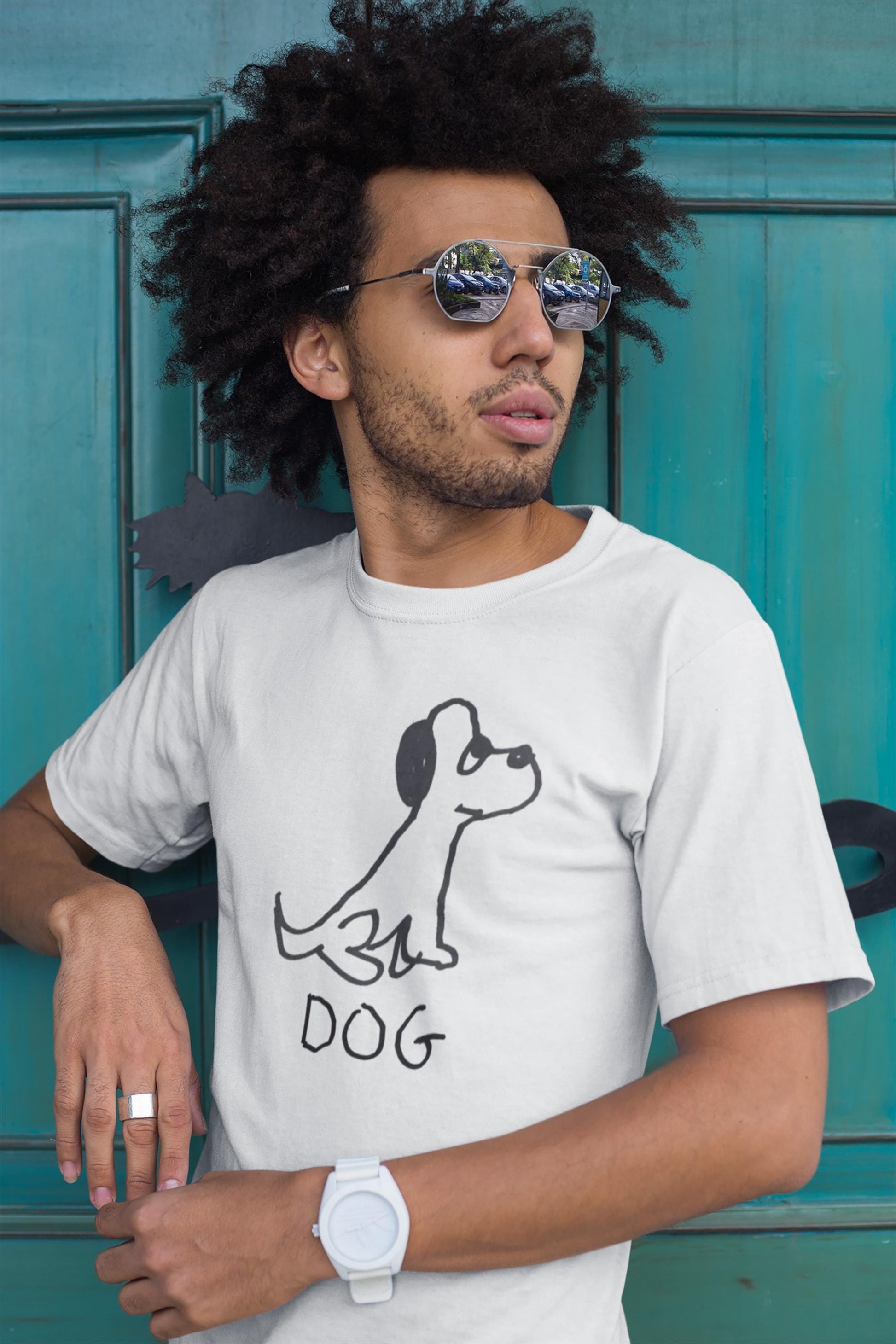 Young man in front of green door wearing a classic white t-shirt featuring a crude but cute black and white marker marker drawing of a cute dog with the word "DOG" handwritten underneath