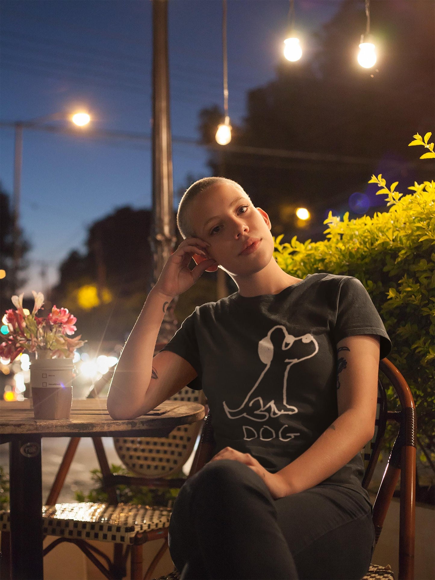 Young gender fluid person sitting at a cafe at night wearing a classic black t-shirt featuring a crude but cute black and white marker marker drawing of a cute dog with the word "DOG" handwritten underneath