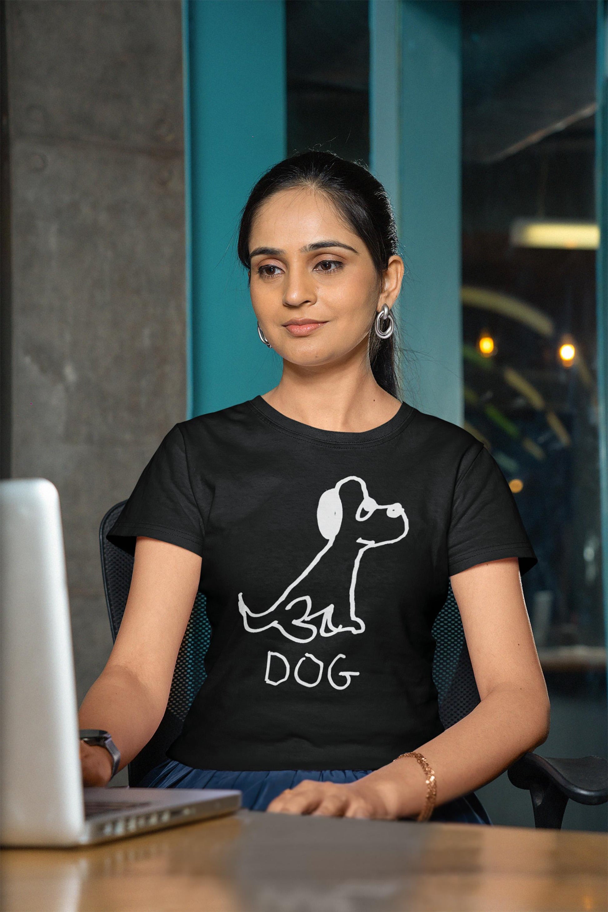 Woman wearing a black t-shirt featuring a crude marker drawing of a dog with the word "DOG" handwritten underneath, sitting at a desk on her laptop computer. 