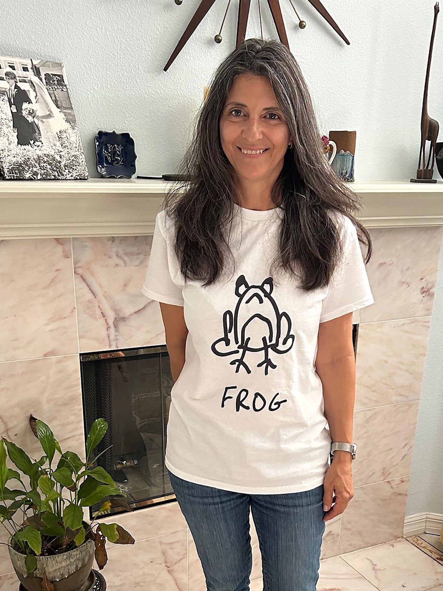 Woman wearing a white t-shirt featuring a crude but cute black and white marker marker drawing of a cute frog with the word “FROG” handwritten underneath, standing in a room with a fireplace.