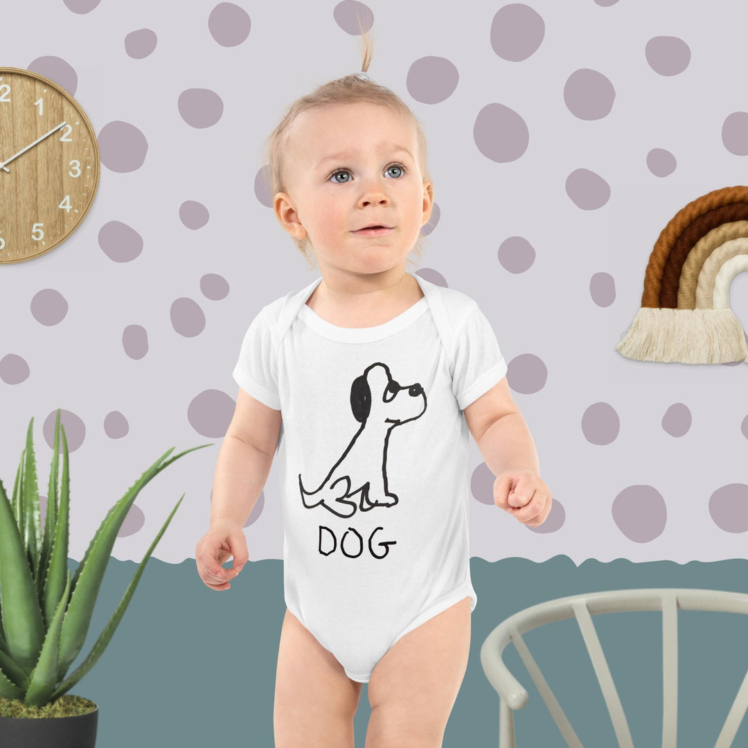 Baby wearing a white onesie featuring a crude marker black and white marker drawing of a cute dog with the word "DOG" handwritten underneath, standing against a polka dot wall.