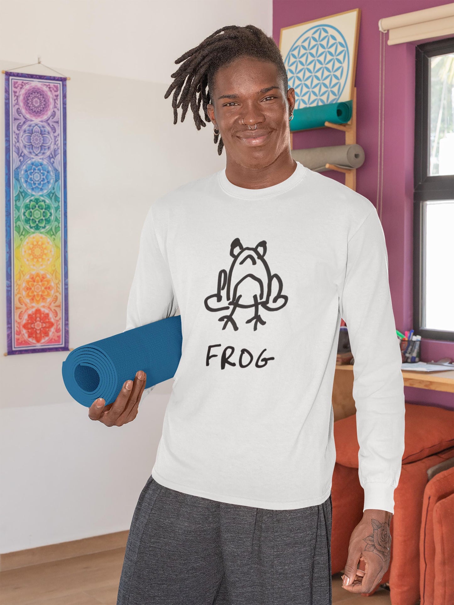 Man carrying a yoga mat wearing a white long sleeved tee shirt featuring a crude but cute black and white marker marker drawing of a cute frog with the word “FROG” handwritten underneath