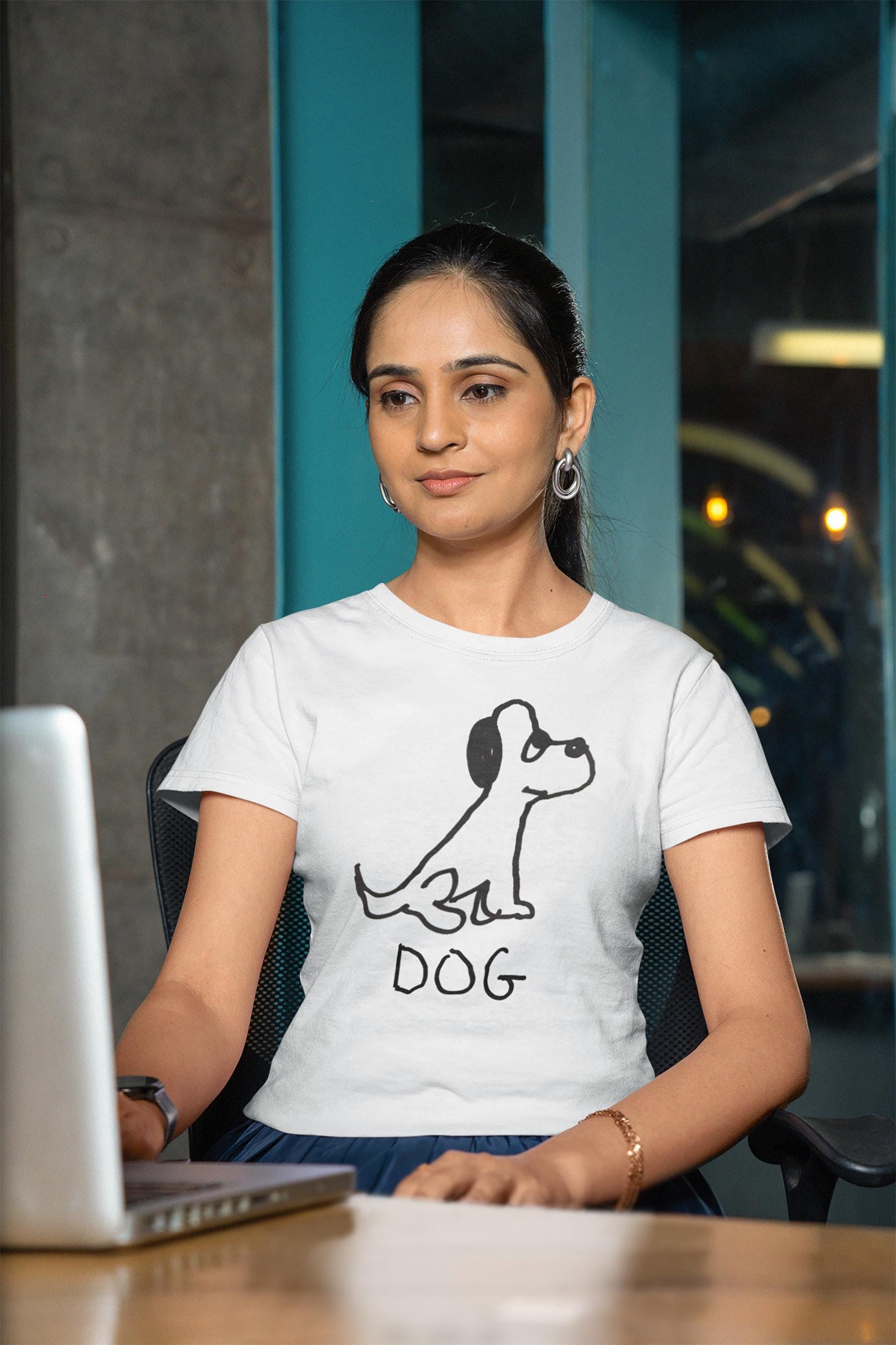 Woman wearing a white t-shirt featuring a crude marker drawing of a dog with the word "DOG" handwritten underneath, sitting at a desk on her laptop computer. 