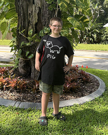 Child wearing a black t-shirt with a hand-drawn lo-fi Pig graphic and text, standing outdoors near a tree and garden.
